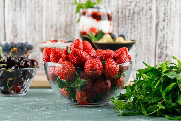 Dessert in vaso e calice con fragole, mirtilli, menta, vista laterale di ciliegie su gesso e superficie sgangherata