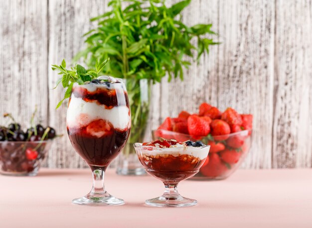 Dessert in vaso e calice con fragole, mirtilli, menta, vista laterale di ciliegie su superficie rosa e sgangherata