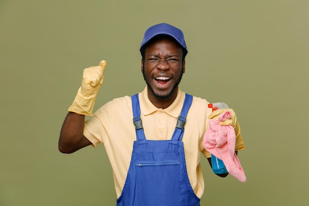 Detergente per la tenuta eccitato con straccio giovane maschio pulitore afroamericano in uniforme con guanti isolati su sfondo verde