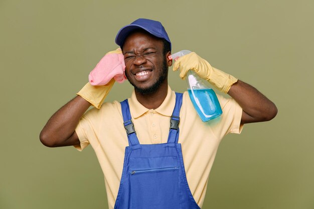 Detergente per la tenuta eccitato con straccio giovane maschio pulitore afroamericano in uniforme con guanti isolati su sfondo verde