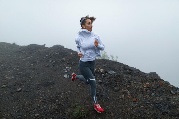 Determinata corridore femminile che fa jogging su una scogliera con tempo nebbioso freddo e ascolta musica con gli auricolari Copia spazio