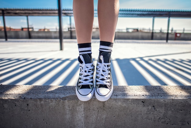 Dettagli delle gambe di donna che si levano in piedi sul bordo di calcestruzzo nelle sue classiche scarpe da ginnastica