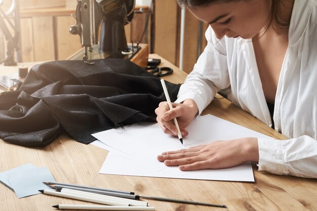 Devo scriverlo fino a quando non mi è sfuggito di mente. Stilista creativa focalizzata seduta in officina e disegnando un nuovo progetto di indumento che cucirà sulla macchina per cucire. Il primo è il piano successivo: l'azione