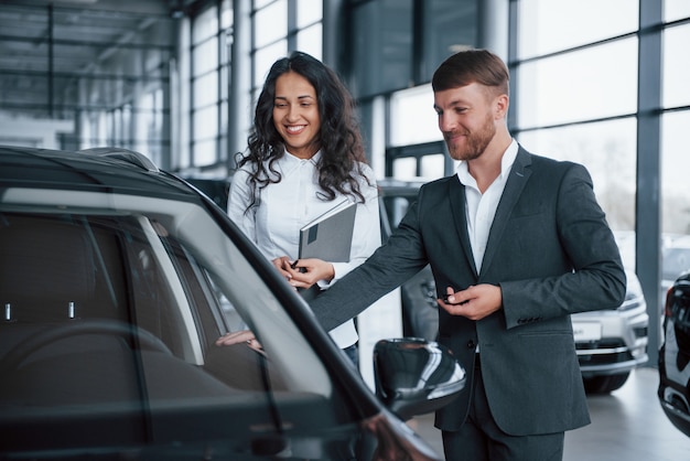 Di buon umore. Cliente femminile e uomo d'affari barbuto elegante moderno nel salone dell'automobile