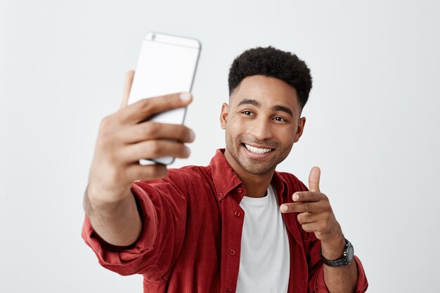 Di 'Cheese. Chiuda su di giovane bello uomo dalla carnagione scura con l'acconciatura afro in maglietta bianca casuale e la camicia rossa che sorridono con i denti, tenendo lo smartphone, facendo la foto del selfie.