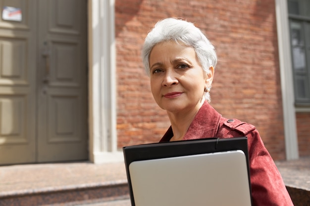 di donna di mezza età moderna impegnata con i capelli grigi in posa all'esterno di un edificio in mattoni, che trasportano gadget digitali alla moda per il lavoro a distanza o l'istruzione online