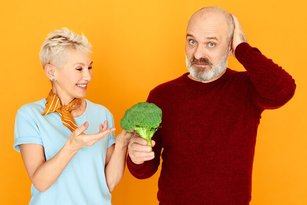 Dieta, cibo, salute, prodotti biologici e concetto di vegetarismo. Uomo anziano frustrato che guarda l'obbiettivo con espressione facciale triste, tenendo broccoli disgustosi, sua moglie gli fa mangiare verdure