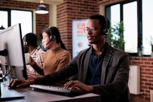 Dipendente afroamericano della linea di assistenza che lavora alla reception del call center con più monitor. Operatore maschio che utilizza le telecomunicazioni per aiutare i clienti al supporto del servizio clienti, rete remota.