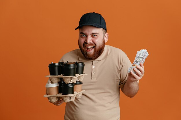 Dipendente dell'uomo delle consegne in berretto nero e uniforme della maglietta vuota che tiene tazze di caffè e contanti che sembra felice ed eccitato che sorride allegramente in piedi su sfondo arancione