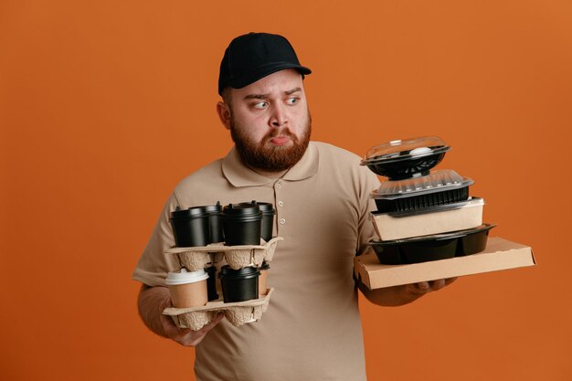 Dipendente dell'uomo delle consegne in berretto nero e uniforme della maglietta vuota che tiene tazze di caffè e contenitori per alimenti guardando confuso le guance che soffiano avendo dubbi in piedi su sfondo arancione