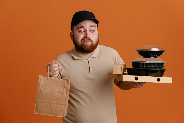 Dipendente dell'uomo delle consegne in berretto nero e uniforme di maglietta vuota che tiene contenitori per alimenti con sacchetto di carta che guarda da parte con espressione scettica in piedi su sfondo arancione