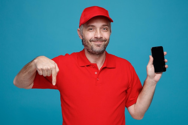 Dipendente dell'uomo delle consegne in uniforme della maglietta bianca con cappuccio rosso che tiene lo smartphone puntato con il dito indice verso il basso guardando la fotocamera sorridente in piedi su sfondo blu