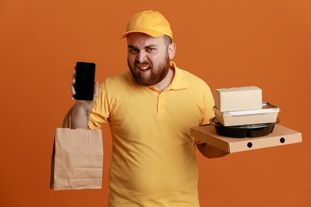 Dipendente dell'uomo delle consegne in uniforme della maglietta vuota con cappuccio giallo che tiene contenitore per alimenti e scatola per pizza con sacchetto di carta che mostra lo smartphone infastidito in piedi su sfondo arancione