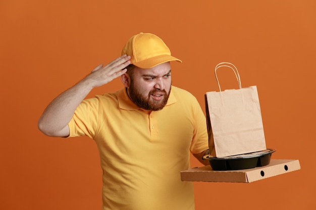 Dipendente dell'uomo delle consegne in uniforme della maglietta vuota con cappuccio giallo che tiene contenitore per alimenti e scatola per pizza con sacchetto di carta che sembra confuso tenendo la mano sulla testa per errore in piedi su sfondo arancione