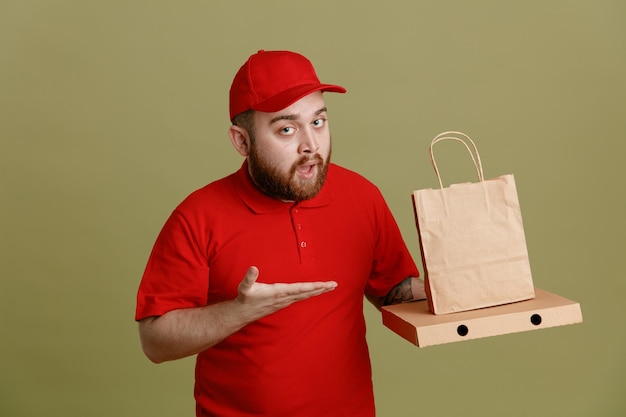 Dipendente dell'uomo delle consegne in uniforme della maglietta vuota con cappuccio rosso che tiene la scatola della pizza e il sacchetto di carta che si presenta con il braccio della mano che sembra sicuro in piedi su sfondo verde