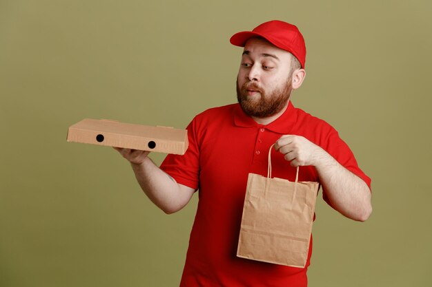 Dipendente dell'uomo delle consegne in uniforme della maglietta vuota con cappuccio rosso che tiene la scatola della pizza e il sacchetto di carta guardando la scatola che è confusa e sorpresa in piedi su sfondo verde