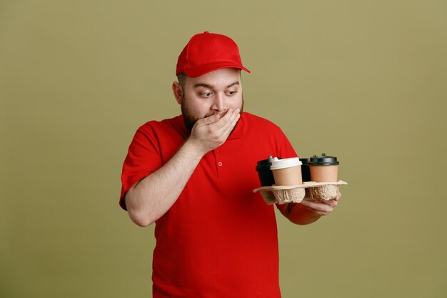 Dipendente dell'uomo delle consegne in uniforme della maglietta vuota con cappuccio rosso che tiene le tazze di caffè guardandole stupito e sorpreso che copre la bocca con la mano in piedi su sfondo verde