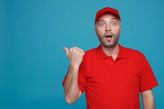 Dipendente dell'uomo di consegna in uniforme della maglietta vuota del cappuccio rosso che esamina la macchina fotografica stupito e sorpreso che indica con il pollice al lato che si leva in piedi sopra fondo blu