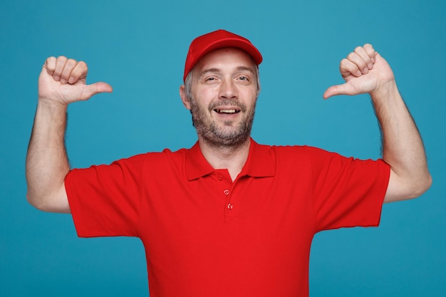 Dipendente dell'uomo di consegna in uniforme della maglietta vuota del cappuccio rosso che guarda la macchina fotografica sorridente soddisfatto di sé che indica se stesso in piedi su sfondo blu