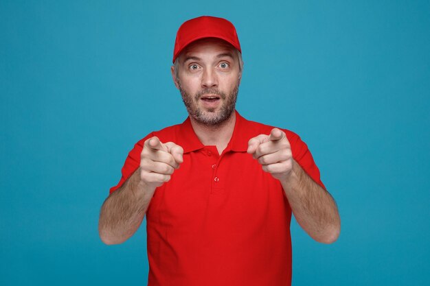 Dipendente dell'uomo di consegna in uniforme della maglietta vuota del cappuccio rosso che guarda la macchina fotografica stupito e sorpreso che indica con il dito indice con entrambe le mani alla macchina fotografica che si leva in piedi sopra priorità bassa blu