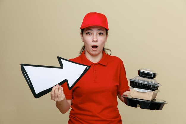 Dipendente donna di consegna in berretto rosso e maglietta vuota uniforme che tiene contenitori per alimenti e grande freccia guardando la fotocamera stupito e sorpreso in piedi su sfondo marrone