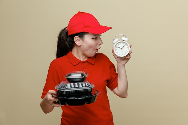 Dipendente donna di consegna in berretto rosso e maglietta vuota uniforme che tiene contenitori per alimenti e sveglia guardandolo stupito e sorpreso in piedi su sfondo marrone