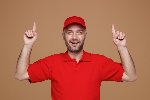 Dipendente uomo di consegna in uniforme tshirt bianca con cappuccio rosso che punta con le dita indice in alto sorridendo fiducioso guardando la fotocamera in piedi su sfondo marrone