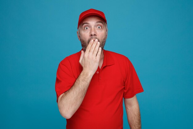 Dipendente uomo di consegna in uniforme tshirt bianca con cappuccio rosso guardando la fotocamera scioccata che copre la bocca con la mano in piedi su sfondo blu