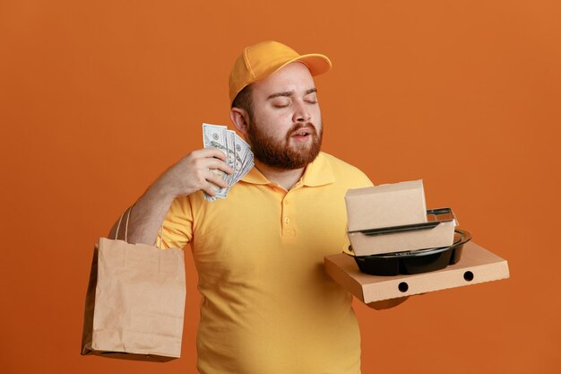 Dipendente uomo di consegna in uniforme tshirt vuota con cappuccio giallo che tiene contenitore per alimenti e scatola per pizza con sacchetto di carta che tiene contanti felice e soddisfatto in piedi su sfondo arancione