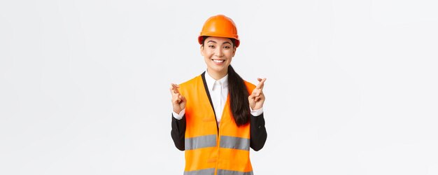 Direttore della costruzione dell'ingegnere femminile asiatico sorridente speranzoso e ottimista nel casco di sicurezza che ha fede nel progetto incrocia le dita buona fortuna e sembra positivo in piedi sullo sfondo bianco