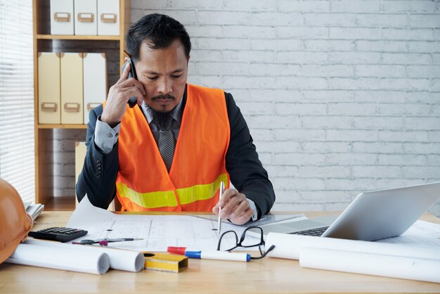 Dirigente maschio asiatico della ditta di costruzioni che si siede nell'ufficio e che parla sul telefono