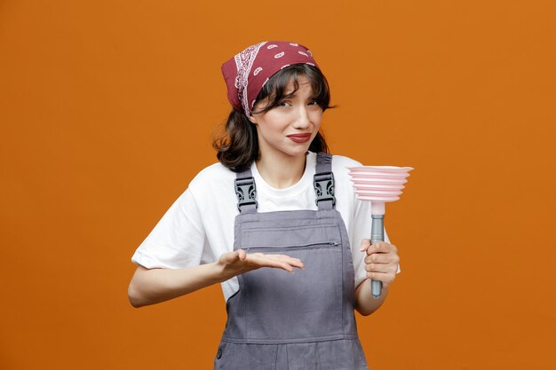 Disgustato giovane pulitore femminile che indossa uniforme e bandana tenendo lo stantuffo che punta verso di esso con la mano che guarda la fotocamera isolata su sfondo arancione