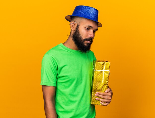 Dispiaciuto che guarda in basso il giovane ragazzo afroamericano che indossa un cappello da festa e tiene in mano una scatola regalo