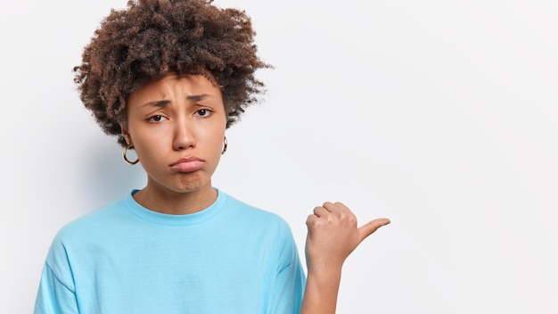 Dispiaciuto giovane dai capelli ricci miserabile giovane donna con i capelli afro si lamenta di qualcosa esprime antipatia mostra brutta cosa punti pollice lontano sullo spazio copia per banner vestito con maglietta blu.