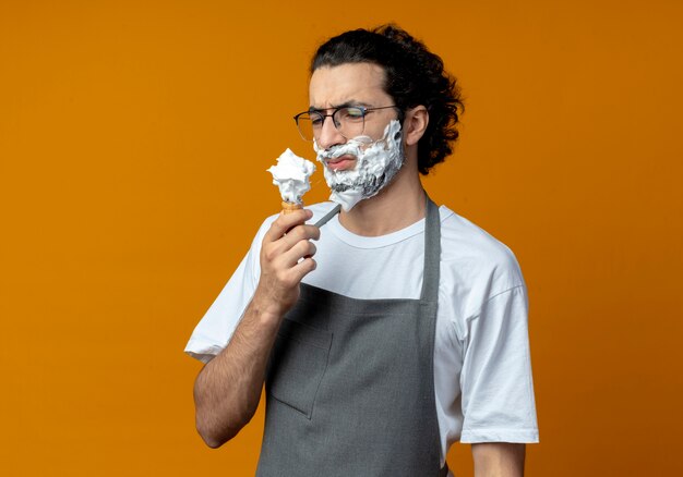 Dispiaciuto giovane maschio caucasico barbiere con gli occhiali e fascia per capelli ondulati in uniforme tenendo e guardando il pennello da barba con crema da barba messo sulla barba