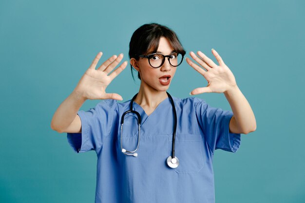 Dispiaciuto mano alzata giovane dottoressa che indossa uno stetoscopio fitth uniforme isolato su sfondo blu