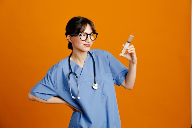 Dispiaciuto mettere la mano sui fianchi tenendo la siringa giovane dottoressa che indossa uno stetoscopio fith uniforme isolato su sfondo arancione