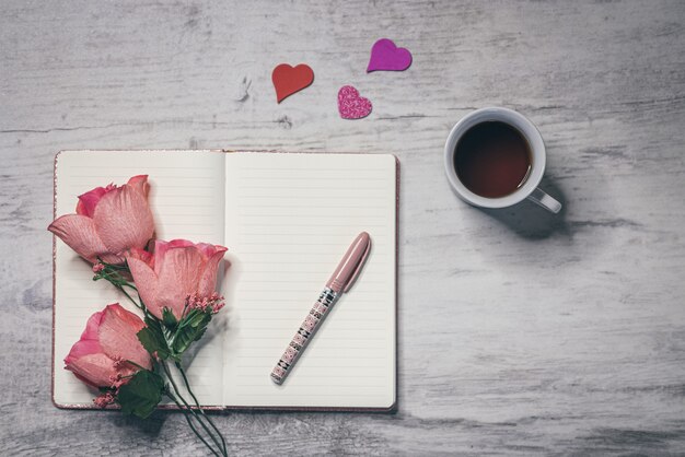 Disposizione piatta di una tazza di caffè, fiori rosa tenui e una penna sulla superficie aperta del taccuino