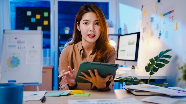 Distanziamento sociale della donna d'affari asiatica nella nuova normalità per la prevenzione dei virus guardando la presentazione della fotocamera al collega sul piano in videochiamata mentre si lavora nella notte dell'ufficio.