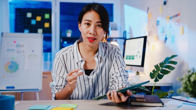 Distanziamento sociale della donna d'affari asiatica nella nuova normalità per la prevenzione dei virus guardando la presentazione della fotocamera al collega sul piano in videochiamata mentre si lavora nella notte dell'ufficio.