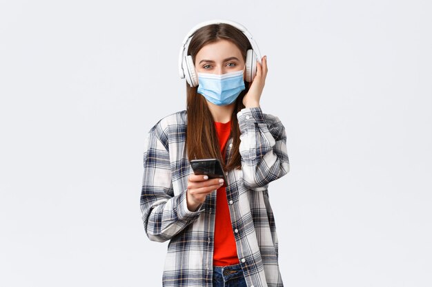 Distanziamento sociale, tempo libero e stile di vita sull'epidemia di covid-19, concetto di coronavirus. Donna sorridente allegra in maschera medica, che sceglie la canzone dallo smartphone della playlist, ascolta la musica con le cuffie wireless.