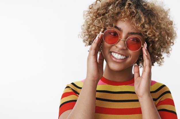 Diva della moda pronta per la passeggiata. Bella ragazza afroamericana elegante e felice con capelli biondi ricci in occhiali da sole rotondi alla moda inclinando la testa toccando i fotogrammi e sorridendo ampiamente alla telecamera