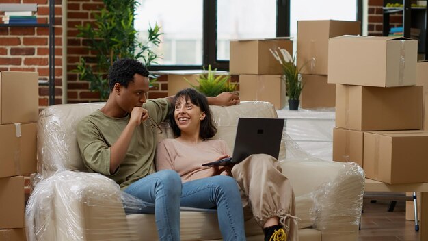 Diverse coppie che navigano in Internet per acquistare decorazioni su laptop, acquistare mobili per la casa su acquisti online per decorare un appartamento soggiorno. Muoversi insieme in una casa in affitto. Scatto da treppiede.