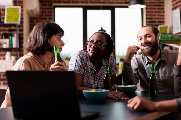 Diversi amici positivi a casa che ridono e parlano insieme mentre si guardano film sul laptop. Persone sorridenti felici in soggiorno che si godono attività ricreative divertenti mentre si mangiano spuntini.