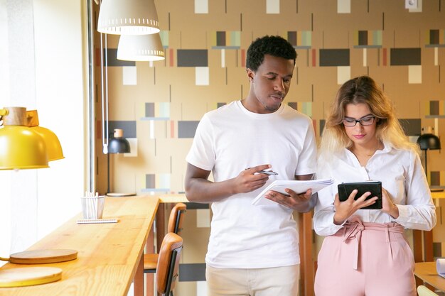 Diversi colleghi che discutono del progetto in ufficio di coworking, utilizzando tablet insieme, scrivendo note in notebook mentre si è in piedi nei luoghi di lavoro e parlando