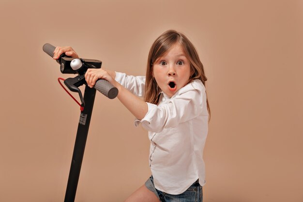 Divertente adorabile bambina indossa una camicia bianca con scooter elettrico in posa con emozioni di sorpresa