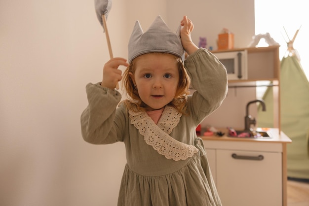 Divertente bambina dalla pelle chiara in abito indossa una corona giocattolo e una bacchetta magica in camera Concetto di bambino