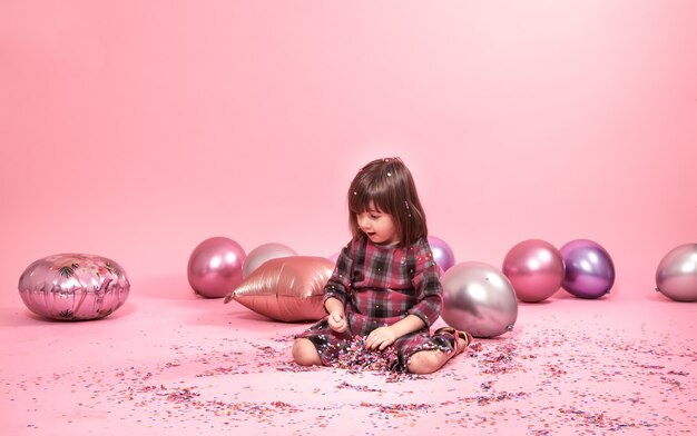 Divertente bambino seduto su uno sfondo rosa. Bambina che si diverte con palloncini e coriandoli