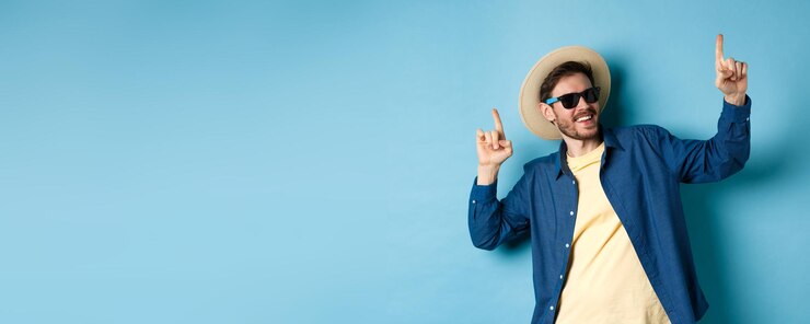 Foto gratuito divertente danza turistica in vacanza puntando le dita verso l'alto indossando cappello estivo e occhiali da sole backgro blu