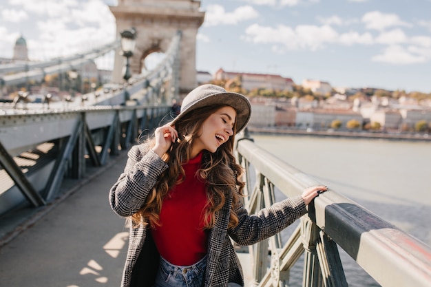 Divertente donna dai capelli lunghi in cappello in posa con gli occhi chiusi durante il servizio fotografico sul ponte in una giornata di sole